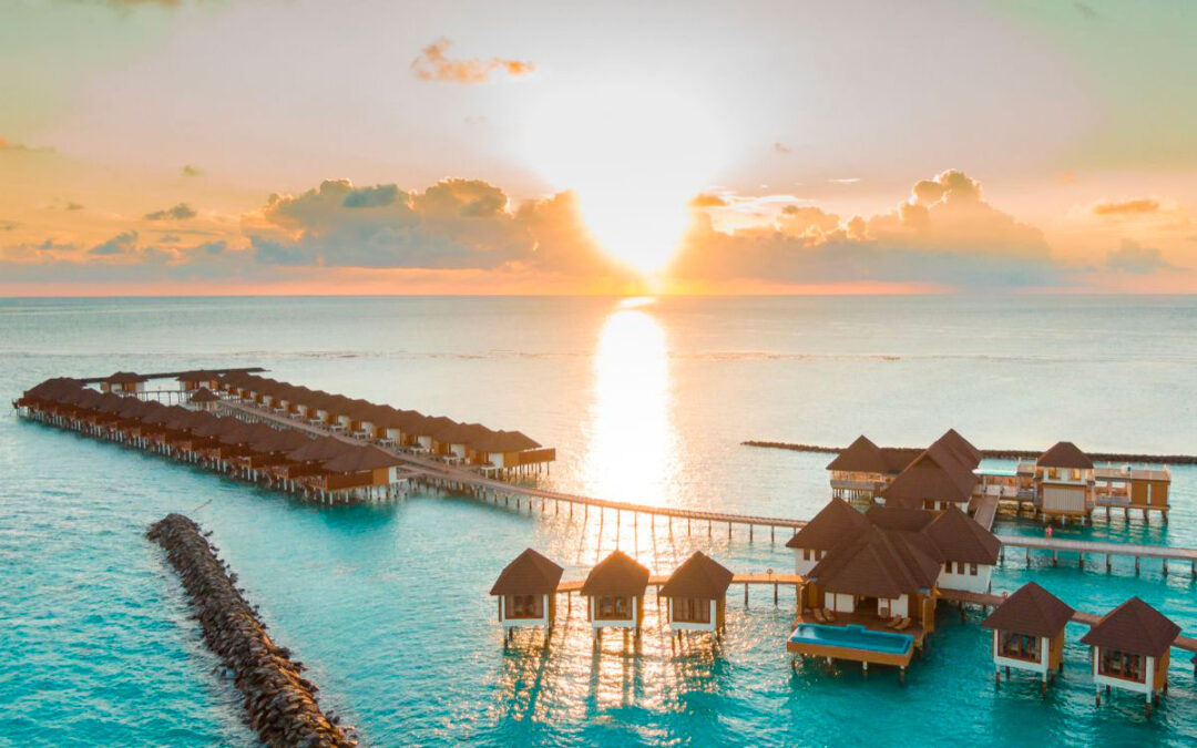 Villas sur pilotis aux Maldives au coucher du soleil, avec une vue panoramique sur l'océan, illuminées par les derniers rayons dorés du soleil, reflétant des teintes chaudes et apaisantes sur les eaux turquoise.